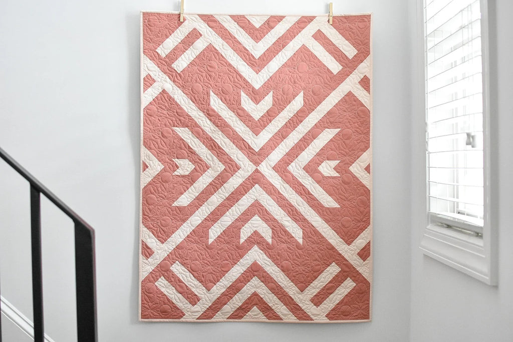 Pink on pink geometric patterned quilt hanging on a wall. Homecoming 2 color quilt kit designed by Lo & Behold Stitchery.